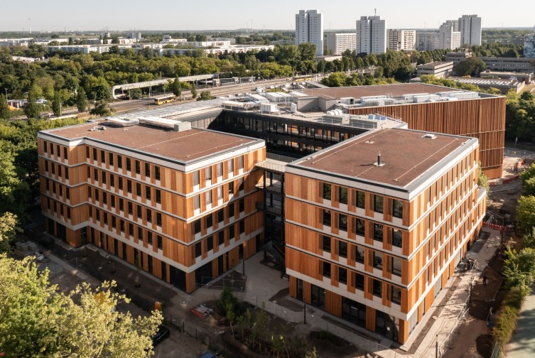 Holzverkleidetes Schulgebäude aus der Vogelperspektive. Im Hintergrund sind Bäume und hohe Wohngebäude zu erkennen.