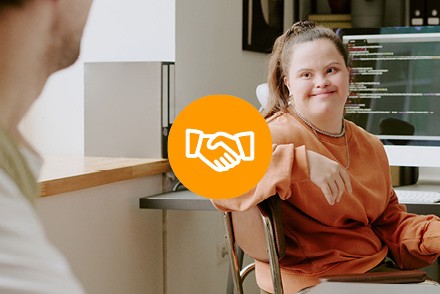 Eine junge Frau schaut lächelnd zu einer anderen Person. Im Hintergrund ist ein Computerbildschirm zu sehen. In der Mitte des Bildes befindet sich in einem orangen Kreis ein Icon, das einen Handschlag darstellt.