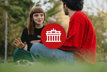 Auf einer Wiese sitzen zwei Personen und unterhalten sich. In der Mitte befindet sich ein roter Kreis mit einem Icon, das ein Gebäude darstellt.