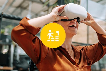 Eine Frau schaut lachend durch eine VR-Brille. In der Mitte des Bildes ist ein gelber Kreis mit einem Icon zu sehen, auf dem eine Person Stufen zu einer Zielfahne hinaufläuft.